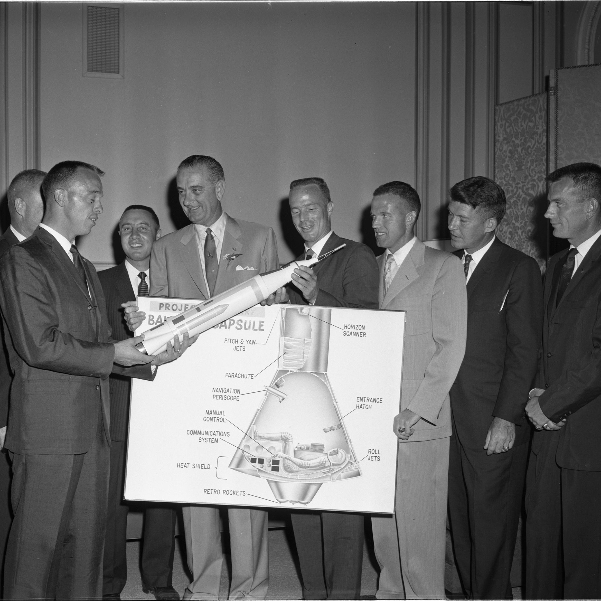 A group of men in suits hold a model rocket and a diagram of a space capsule. 